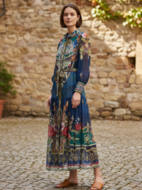 MOSSÉE patterned long‑sleeve maxi dress with vibrant jewel‑tone prints, worn outdoors against a rustic stone wall.